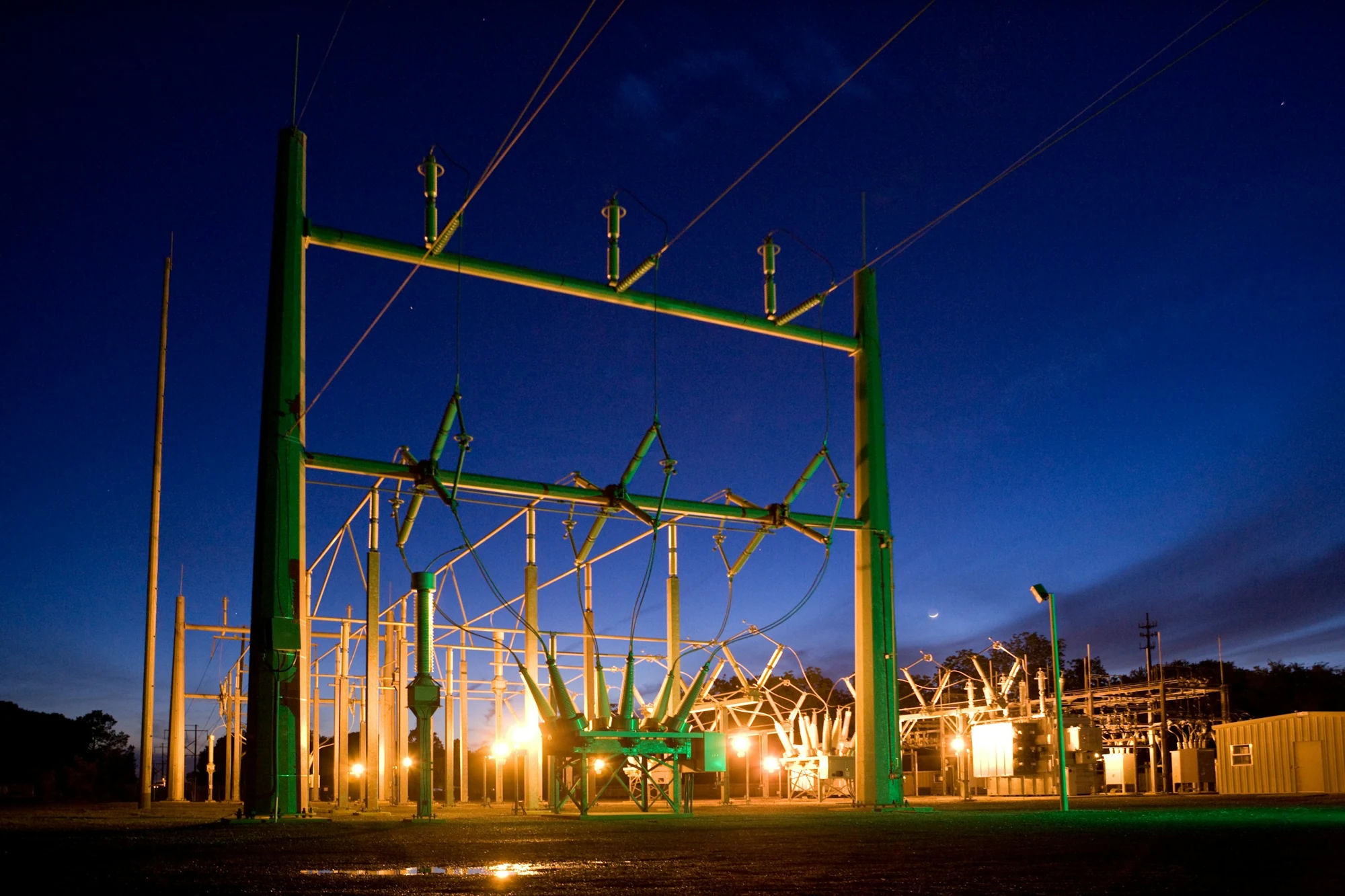 An illuminated public-power substation and transmission equipment at blue hour.